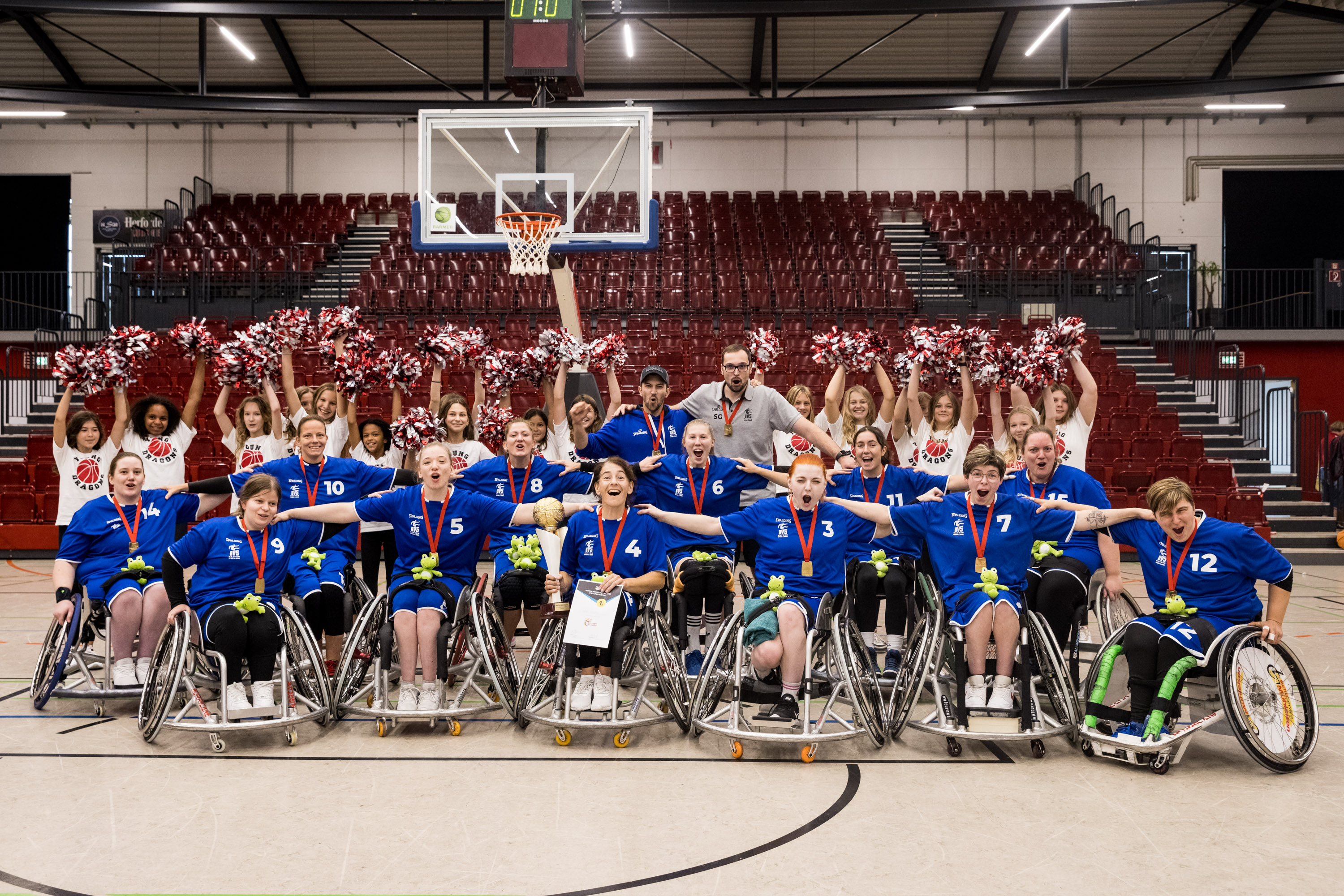Das bayerische Team der Damen im Rollstuhlbasketball hat sich zu einem Gruppenfoto in der Halle aufgestellt. Sie tragen Goldmedaillen, hinter ihnen sieht man Pompons.