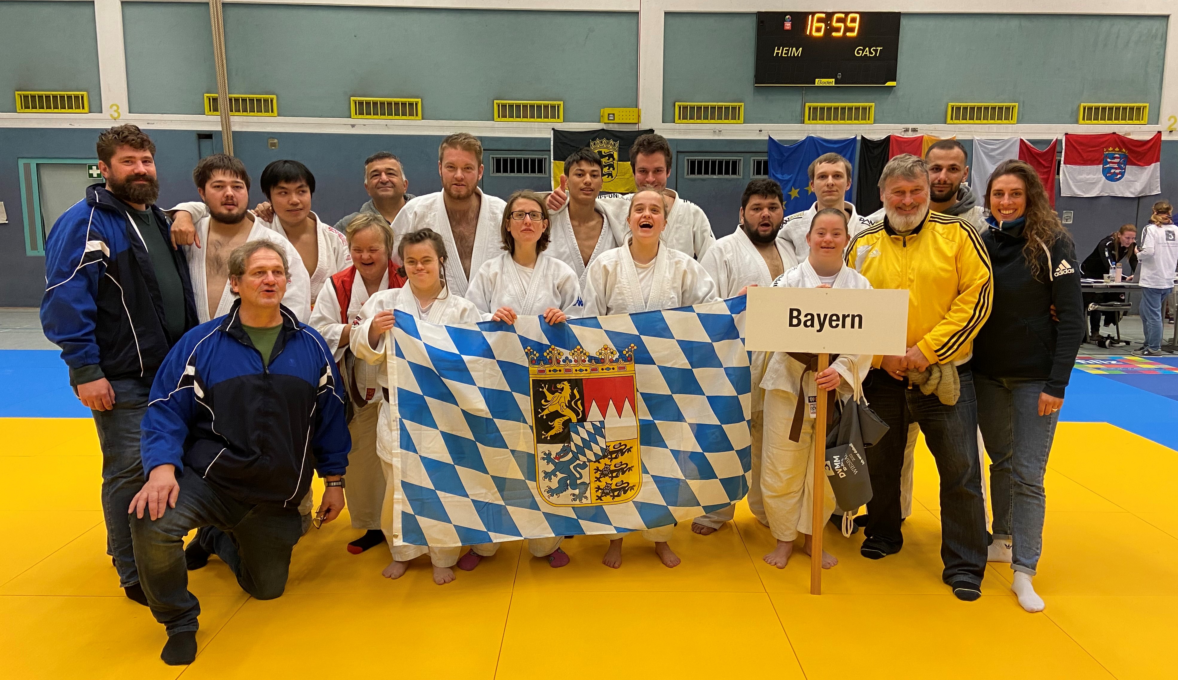 Unsere Judoka posieren in der Sporthalle für ein Gruppenbild und halten eine bayerische Flagge.