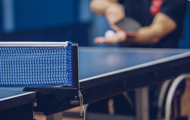 Ein Athlet im Para Tischtennis sitzt an einer Platte und schlägt auf. Im Vordergrund sieht man das Netz