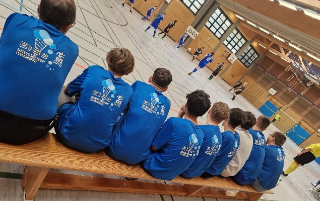 Sportler*innen mit blauen T-Shirts sitzen auf einer Langbank in einer Halle und schauen sich ein Fußballspiel an