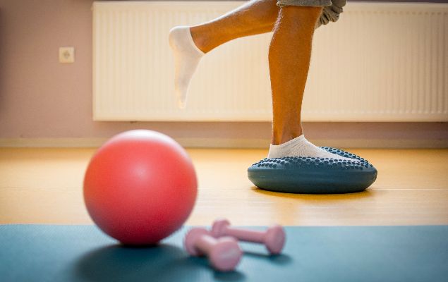 Ein Gymastikball und Kurzhanteln liegen auf einer Sportmatte. Im Hintergrund wird eine Sportübung ausgeführt.