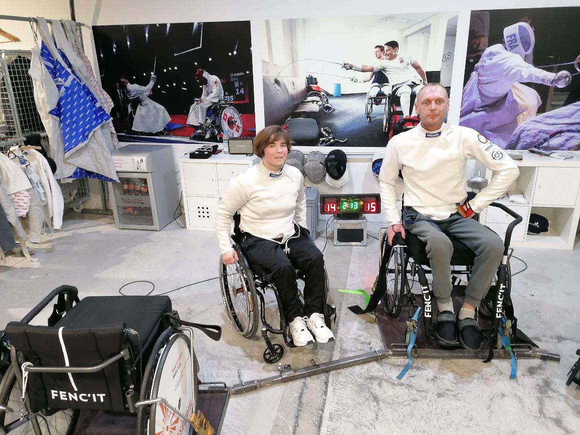 Eine Rollstuhlfechterin und ein Rollstuhlfechter ohne Visier, die in die Kamera blicken. Im Hintergrund hängen verschiedene, große Bilder mit Rollstuhlfecht-Szenen an der Wand. 
