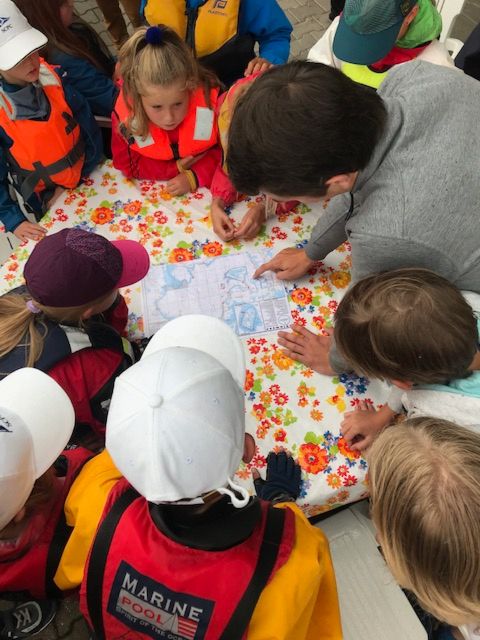 Eine Seekarte des Chiemsees liegt auf einem Tisch. Kinder und Jugendliche stehen außenherum, ein Trainer zeigt etwas auf der Karte. 