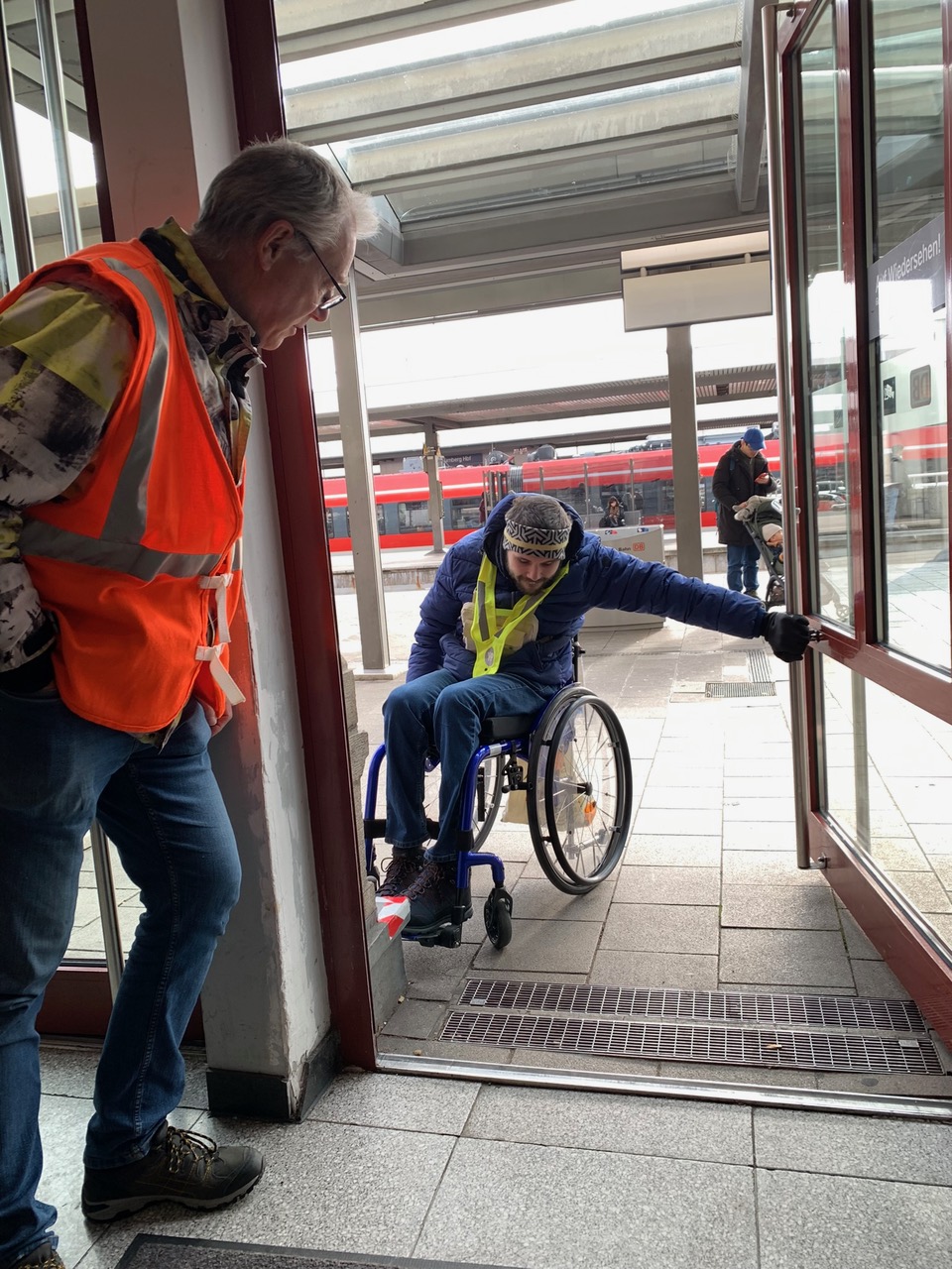 Mann im Rollstuhl öffnet mit der Hand eine große Türe am Bahnhof.