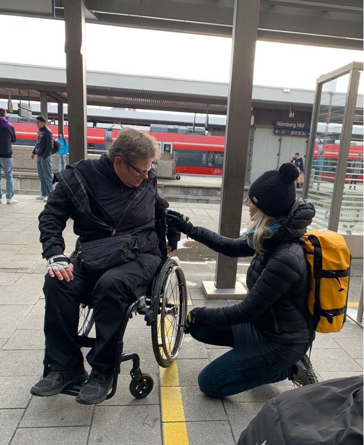 Mann im Rollstuhl am Bahnsteig, neben ihm eine hockende Frau mit Hand am Rollstuhl