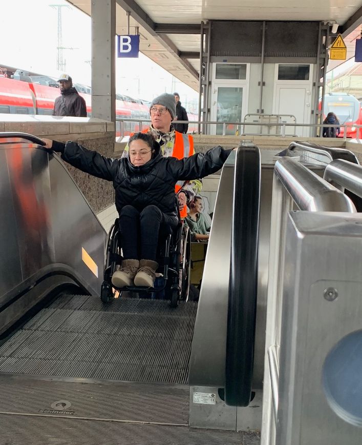 Frau im Rollstuhl auf Rolltreppe am Bahnhof.