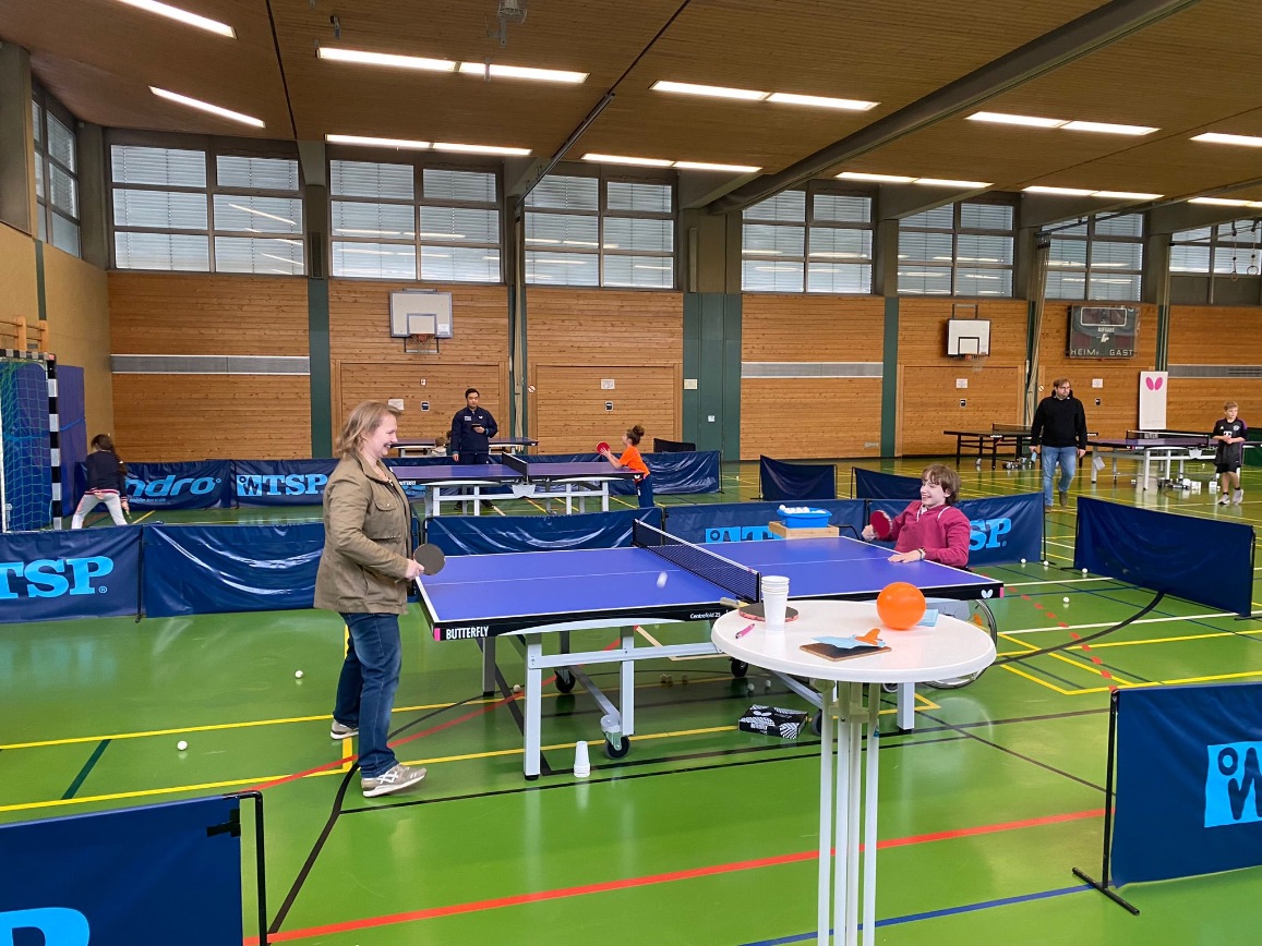 Junge im Rollstuhl und Fußgängerin spielen Tischtennis in einer Sporthalle